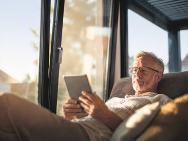 Mann sitzt mit Tablet auf dem Sofa und stellt die Öffnung der Fenster über smart home ein