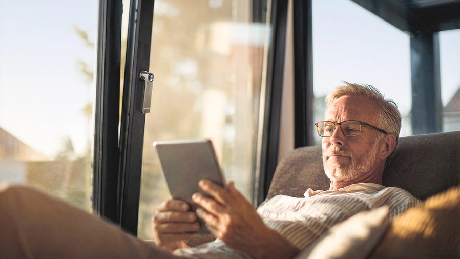 Mann sitzt mit Tablet auf dem Sofa und stellt die Öffnung der Fenster über smart home ein