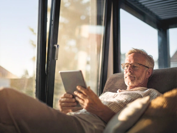 Mann sitzt mit Tablet auf dem Sofa und stellt die Öffnung der Fenster über smart home ein